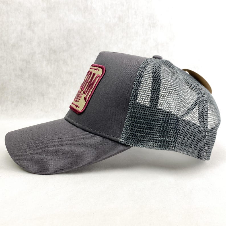 K&ouml;nnte beinhalten: Graue Trucker-Cap mit Mesh-R&uuml;ckseite und gebogenem Schirm. Auf der Vorderseite befindet sich ein rechteckiger Aufn&auml;her mit dem Wort "Ranch" in bordeauxfarbenen Buchstaben. Ein braunes Etikett ist an der R&uuml;ckseite befestigt.