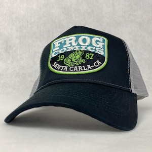Puede incluir: Gorra de camionero negra y gris con un parche bordado en verde, azul y blanco que dice "Frog Comics 1987 Santa Carla-CA" y presenta una rana verde.