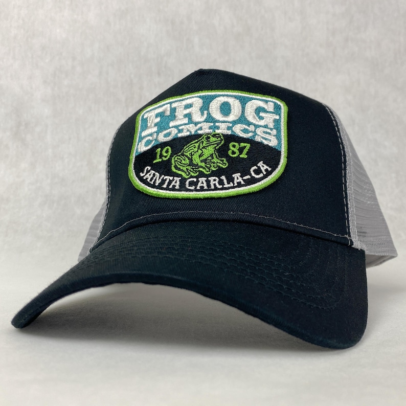 Trucker hat with a black front and white mesh back featuring a green and white patch. The patch reads 'Frog Comics 1987, Santa Carla-CA' with an image of a frog in the center.