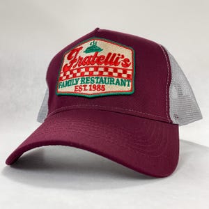 Puede incluir: Una gorra de camionero color burdeos con una malla gris en la parte posterior. La gorra tiene un parche bordado en verde, rojo y blanco que dice "Fratelli's Family Restaurant Est 1985."