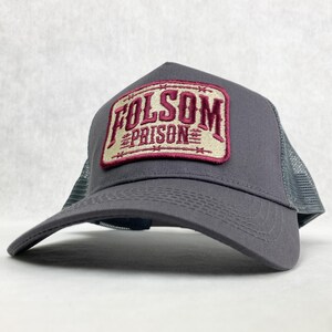 Graphite grey trucker hat with a beige and maroon embroidered patch reading 'Folsom Prison.' The patch has a vintage western style, with barbed wire details around the border. The mesh back adds a classic trucker hat look