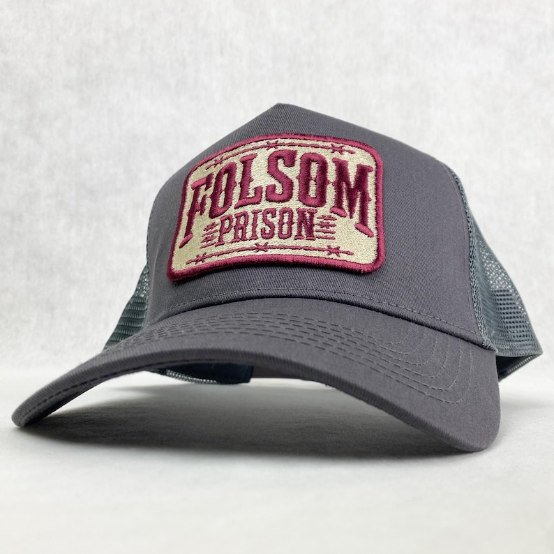 Graphite grey trucker hat with a beige and maroon embroidered patch reading 'Folsom Prison.' The patch has a vintage western style, with barbed wire details around the border. The mesh back adds a classic trucker hat look