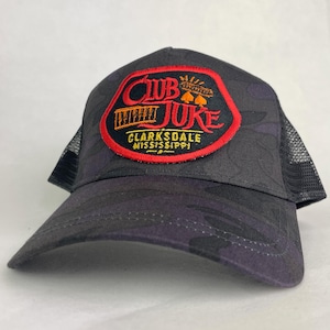 May include: A dark gray camouflage trucker hat featuring a black mesh back and a prominent embroidered patch on the front. The patch is red-bordered and displays the text CLUB JUKE in red and orange, with CLARKSDALE MISSISSIPPI in yellow below.