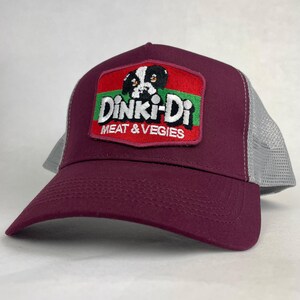 May include: A maroon baseball cap with a grey mesh back and an embroidered patch on the front. The patch features a black and white dog illustration above the text DINKI-DI MEAT & VEGIES in white letters on a red and green background.