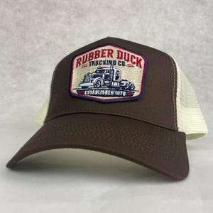 May include: A brown trucker cap with a cream-coloured mesh back. The front panel features a rectangular patch with a blue border, displaying a graphic of a semi-truck and the text RUBBER DUCK TRUCKING CO. ESTABLISHED 1978 in red and blue.