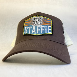 Op de afbeelding: Een bruine truckerpet met een rechthoekige geborduurde patch aan de voorzijde. De patch toont een zwart-witte illustratie van een hond boven het woord STAFFIE in witte letters, geplaatst tegen een blauwe achtergrond met oranje zonnestralen.