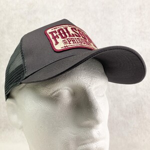 K&ouml;nnte beinhalten: Graue Trucker-Kappe mit gebogenem Schirm und Mesh-R&uuml;ckseite. Die Kappe hat einen rechteckigen Aufn&auml;her mit den Worten "Folsom Prison" in einem weinroten und cremefarbenen Farbschema. Die Kappe wird auf einem Schaufensterkopf pr&auml;sentiert.