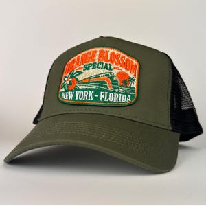 May include: An olive green trucker hat with a black mesh back and a rectangular embroidered patch on the front. The patch features an orange train graphic with the text ORANGE BLOSSOM SPECIAL NEW YORK - FLORIDA in orange and white.