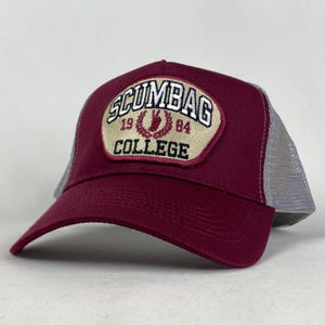 Pode incluir: Boné de caminhoneiro bordô e cinza com o texto "SCUMBAG COLLEGE 1984" bordado em um patch. O boné apresenta uma aba curva e uma parte traseira de malha.