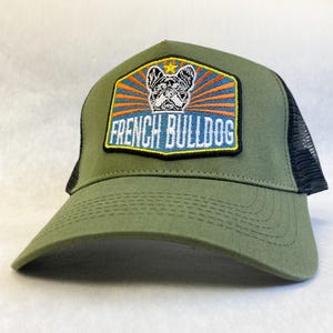 May include: A green and black trucker hat with a patch featuring a cartoon French Bulldog with the text "French Bulldog".