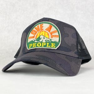 Könnte beinhalten: Schwarze Camouflage-Trucker-Kappe mit einem gestickten Patch in Grün, Orange und Gelb, auf dem "Love All The People" mit einem Berg- und Sonnenmotiv steht.