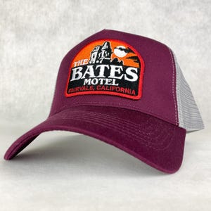 May include: A burgundy baseball cap with a grey mesh back. The front features a patch with the words "THE BATES MOTEL FAIRVALE, CALIFORNIA" and an image of a spooky house and a moon.