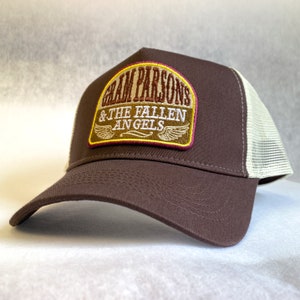 Puede incluir: Una gorra de camionero marrón y blanca con un parche marrón que dice "Gram Parsons & The Fallen Angels" en letras amarillas y blancas con un diseño de alas amarillas y blancas.