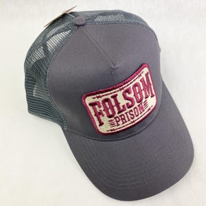 K&ouml;nnte beinhalten: Graue Trucker-Kappe mit Mesh-R&uuml;ckseite und gebogenem Schirm. Die Kappe hat einen rechteckigen Aufn&auml;her mit den Worten "FOLSOM PRISON" in burgunderroter Schrift. Der Aufn&auml;her ist mit einem burgunderroten Rand und einem Stacheldraht-Design versehen.