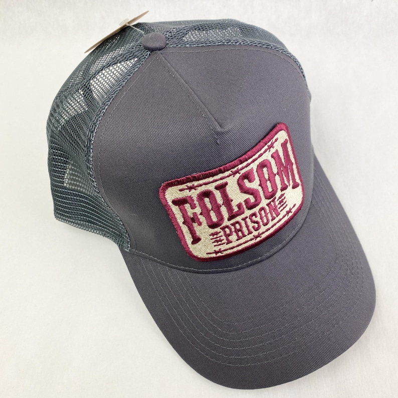 K&ouml;nnte beinhalten: Graue Trucker-Kappe mit Mesh-R&uuml;ckseite und gebogenem Schirm. Die Kappe hat einen rechteckigen Aufn&auml;her mit den Worten "FOLSOM PRISON" in burgunderroter Schrift. Der Aufn&auml;her ist mit einem burgunderroten Rand und einem Stacheldraht-Design versehen.