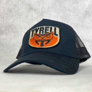 Könnte beinhalten: Schwarze Trucker-Kappe mit einem schwarzen und orangefarbenen gestickten Aufnäher mit einer Eule und dem Text "TYRELL".