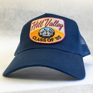 Può includere: Un cappello da camionista blu navy con una toppa rossa, gialla e arancione che dice "Hill Valley Class of '85".