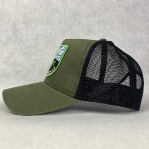 May include: A green and black trucker hat with a green and white embroidered patch that says "Big Sur" on the front. The hat has a black mesh back.