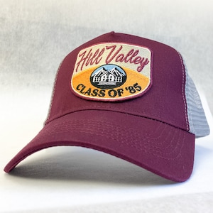 Op de afbeelding: Bordeauxkleurige truckercap met een grijs mesh achterpaneel. De pet heeft een patch met de tekst "Hill Valley Class of '85" en een afbeelding van een gebouw met bergen op de achtergrond.