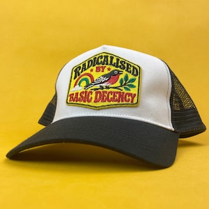 May include: A white and black trucker hat featuring an embroidered patch on the front. The patch displays a bird perched on a branch with a rainbow and the text RADICALISED BY BASIC DECENCY in yellow, red, and black thread on a yellow background.