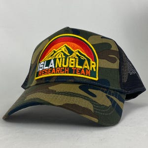 May include: Camouflage baseball cap with a black mesh back. The front has a patch with the text "ISLA NUBLAR RESEARCH TEAM" above a mountain and sunset design.