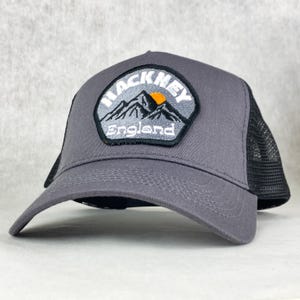 Può includere: Cappellino da camionista grigio con retro in rete nera e visiera curva. La parte anteriore presenta una toppa con la scritta "HACKNEY ENGLAND" sopra un paesaggio montano con un sole giallo. Un accessorio classico.