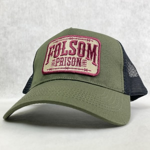 May include: An olive green trucker hat with a black mesh back and a curved brim. The hat features a rectangular patch with the words "FOLSOM PRISON" embroidered in burgundy.
