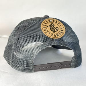 K&ouml;nnte beinhalten: Graue Trucker-Cap mit Mesh-R&uuml;ckseite und gebogenem Schirm. Die Kappe hat einen runden, braunen Lederaufn&auml;her mit den Worten "Trucker Hat Company" und einem Logo. Der verstellbare Riemen ist grau.