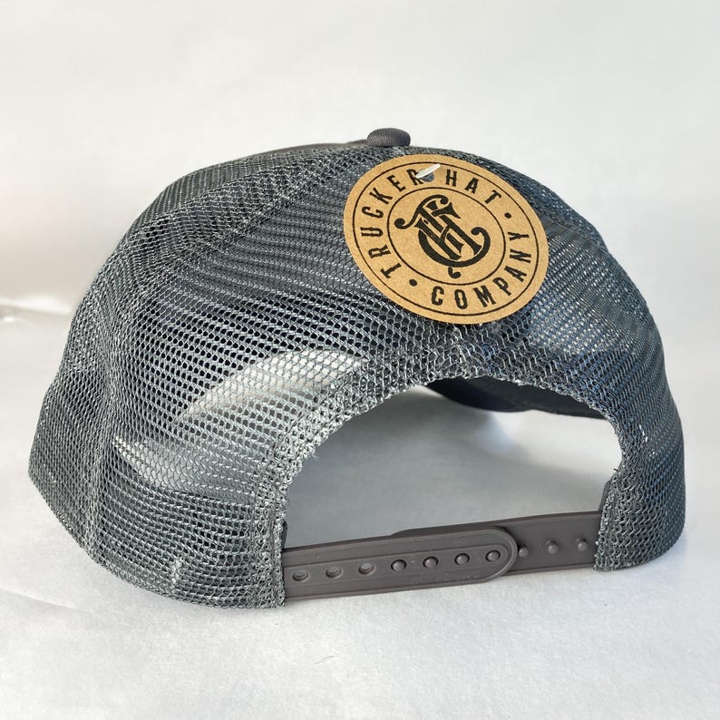 K&ouml;nnte beinhalten: Graue Trucker-Cap mit Mesh-R&uuml;ckseite und gebogenem Schirm. Die Kappe hat einen runden, braunen Lederaufn&auml;her mit den Worten "Trucker Hat Company" und einem Logo. Der verstellbare Riemen ist grau.