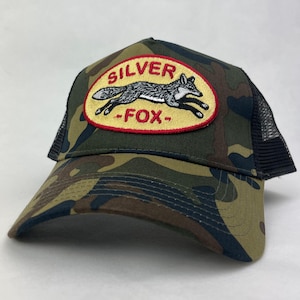 May include: A camouflage trucker hat featuring a black mesh back and a front panel with an oval patch. The patch has a yellow background, a red border, and an embroidered illustration of a running fox with the text SILVER FOX in red letters.