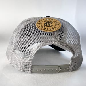 May include: A gray and black trucker hat with a brown patch that says "Trucker Hat Company" with a logo of a stylized "TH" inside a circle.