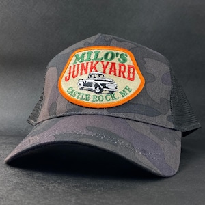 Könnte beinhalten: Eine schwarze und grüne Camouflage-Trucker-Kappe mit einem Patch, auf dem "Milo's Junkyard" mit einem Vintage-Truck-Motiv und dem Text "Castle Rock, ME" steht.