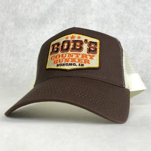 Könnte beinhalten: Eine braune und weiße Trucker-Kappe mit einem Patch, auf dem "BOB'S COUNTRY BUNKER KOKOMO, IN" in orangefarbenen und gelben Buchstaben steht.