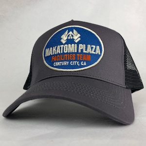 May include: A charcoal grey trucker hat featuring a blue oval patch with white and orange embroidery. The patch displays the text NAKATOMI PLAZA FACILITIES TEAM CENTURY CITY, CA. The cap has a solid front panel and a black mesh back panel.