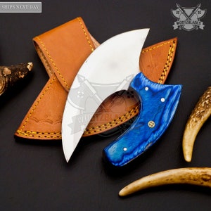 Op de afbeelding: Een blauw en wit gebogen mes met een leren schede. Het mes heeft een houten handvat met een blauw en wit marmerpatroon. De schede is van bruin leer met gele stiksels.