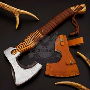 May include: A hand-forged axe with a wood handle and a leather sheath. The axe head has a decorative design and the handle is wrapped in leather. The sheath is brown leather with a decorative stitching pattern. The axe is a traditional design and is perfect for camping, hiking, or survival.