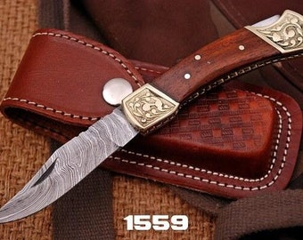 Puede incluir: Un pequeño cuchillo de bolsillo con mango de madera y hoja de acero damasco. El cuchillo tiene una incrustación decorativa de latón en el mango y viene con una funda de cuero marrón. El número 1559 está impreso en la parte inferior de la imagen.