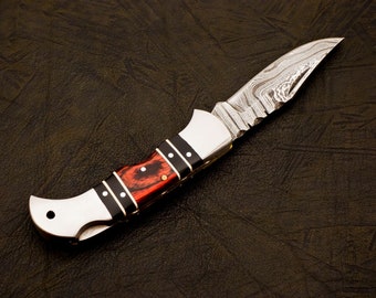 Puede incluir: Un cuchillo plegable de bolsillo con mango de madera negro y rojo y una hoja de acero inoxidable con un patrón de damasco.