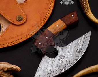 May include: A Damascus steel axe with a wooden handle and a leather sheath. The blade has a unique pattern, and the handle features a combination of light and dark wood. The sheath is brown with yellow stitching.
