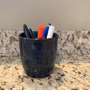 Op de afbeelding: Een zwarte keramische beker met een getextureerd oppervlak bevat een verzameling pennen en stiften, waaronder een blauwe pen, een witte pen en een oranje stift.