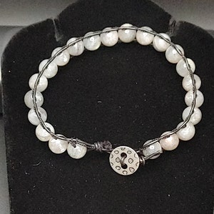 Puede incluir: Un brazalete de piedras blancas con cierre de botón plateado. El brazalete está hecho de cuentas blancas y un cordón negro.