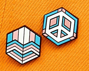 May include: Two enamel pins on a bright orange background. One pin is a white peace sign in a blue and pink geometric shape. The other pin is a geometric shape with pink, blue, and white stripes.
