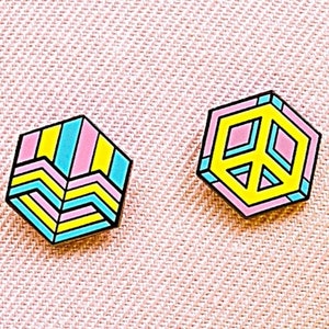 May include: Two colorful enamel pins on a pink fabric background. One pin is a yellow, pink, and blue geometric design. The other pin is a yellow peace sign with a pink outline.