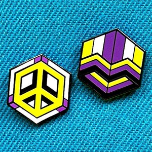 Peut inclure: Deux épingles en émail sur un fond de tissu bleu. Une épingle est un signe de paix jaune avec un contour blanc et des accents violets. L'autre épingle est un motif géométrique violet, jaune et blanc.