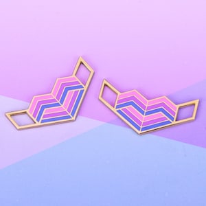 May include: Two gold-colored pins with a geometric design in shades of pink, purple, and blue. The pins are shaped like a heart and are set against a pink and blue background.