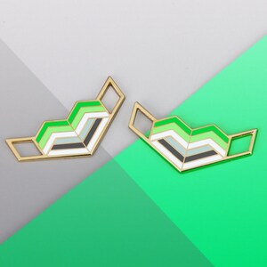 May include: Two gold-colored pins with a geometric design in green, white, and black colors. The pins are shaped like a chevron and are arranged in a symmetrical pattern.