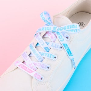 May include: White canvas sneakers with blue and pink shoelaces that have the words "Community", "Identity", "Freedom", "Unity", "Together", "Love", "Kindness", "Hope", "Peace", "Joy", "Compassion", and "Freedom" printed on them.