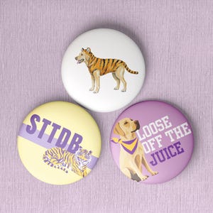 May include: Three circular pin-back buttons. The top button is white with a tiger-striped dog. The bottom left button is pale yellow with a purple diagonal stripe, a tiger, and the text STTDB. The bottom right button is purple with a dog wearing a scarf and the text LOOSE OFF THE JUICE.