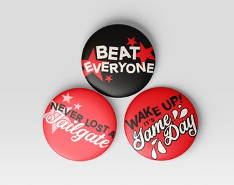 May include: Three round red and black buttons with white text. The first button says "Beat Everyone" with red stars. The second button says "Never Lost A Tailgate" with white stars. The third button says "Wake Up! It's Game Day" with white water drops.
