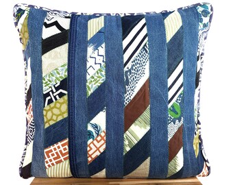 May include: A blue denim pillow with a patchwork design featuring various fabrics in shades of green, brown, white, and orange. The pillow has a square shape and is made with a variety of textures.